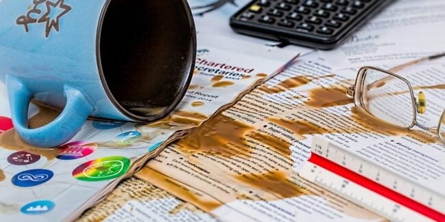 Image of QuickBooks forms spread out, with an overturned coffee cup spilling onto them, illustrating common mistakes that can occur when using QuickBooks and the importance of careful financial management.